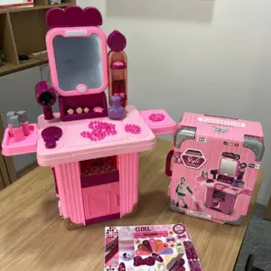 Tocador infantil rosa y magenta con espejo, secador y accesorios, junto a su caja tipo maleta y guía de accesorios sobre una mesa de madera.
